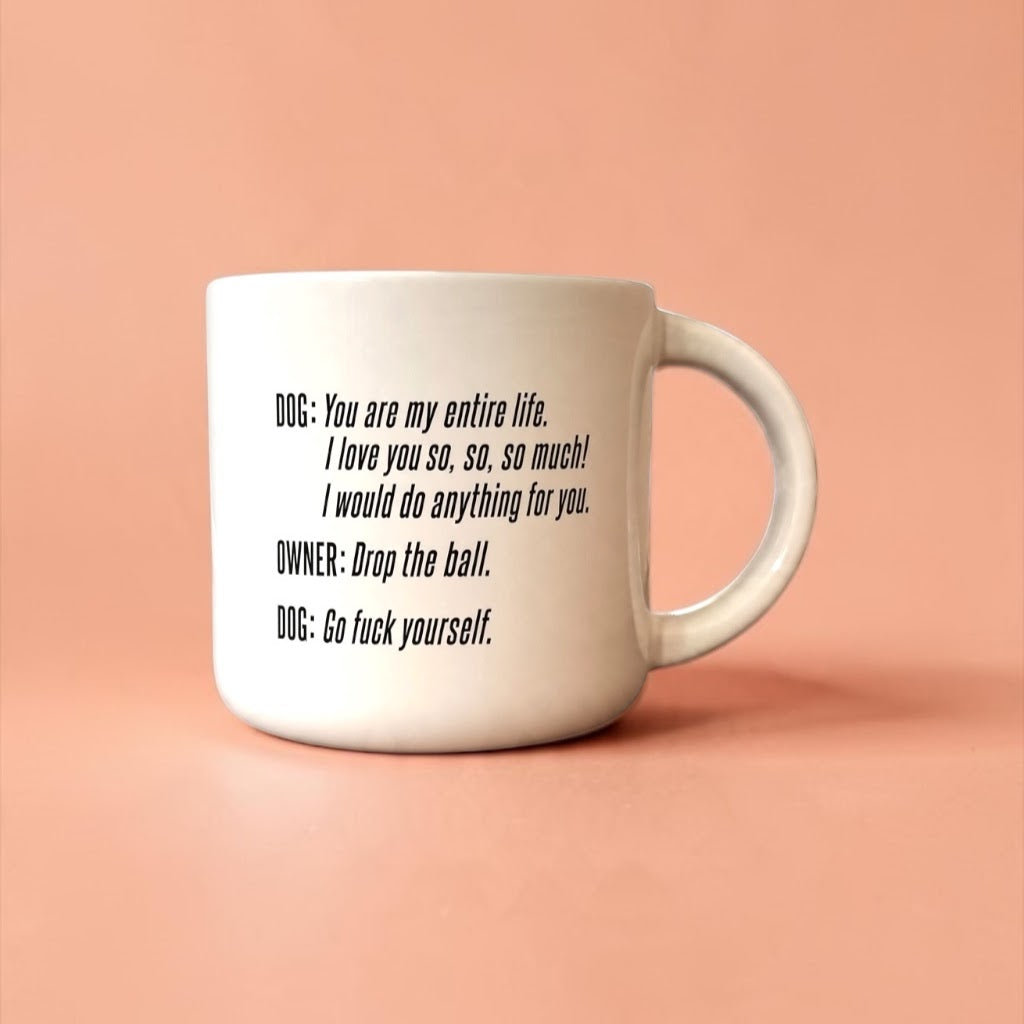 White ceramic coffee mug printed with “DOG: You are my entire life… OWNER: Drop the ball. DOG: Go fuck yourself.” on a peach background.